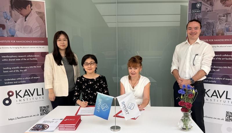 Four people pose at a signing table in front of Kavli Institute for Nanoscience Discovery and University of Oxford banners. Two are seated and signing documents, with small SMART flags and booklets on the table.