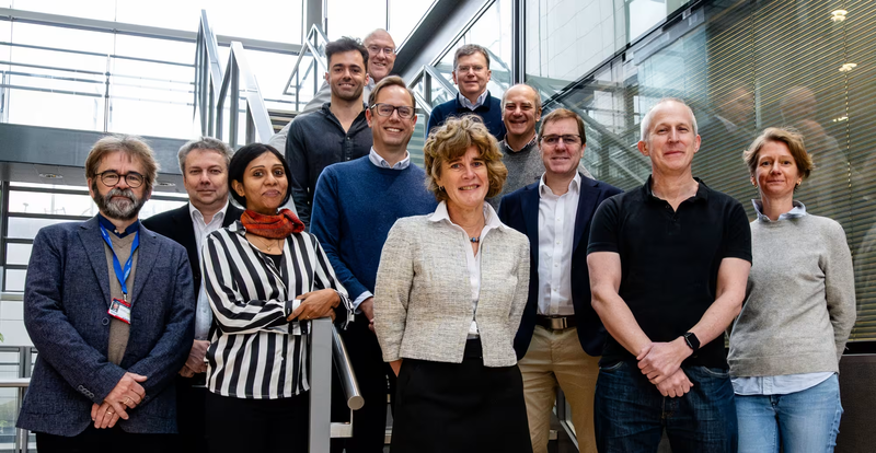 A group of eleven people poses on a staircase inside a modern glass-and-metal building, with several standing in front and others on the steps behind them.