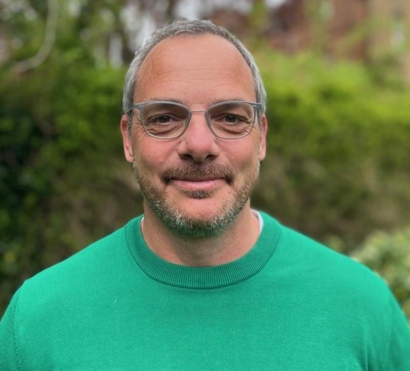 Man with glasses and green jumper smiling into the camera.