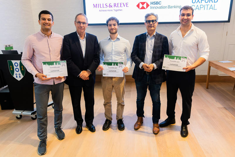Five men pose together indoors, with three holding certificates. A screen behind them displays Mills & Reeve, HSBC Innovation Banking, and Oxford Capital logos.
