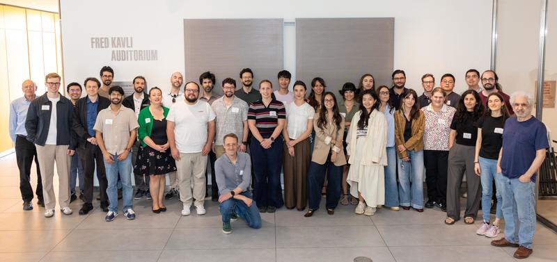 A large group of symposium attendees pose for a group photo inside the Fred Kavli Auditorium, standing in rows in front of a light-colored wall and auditorium signage.