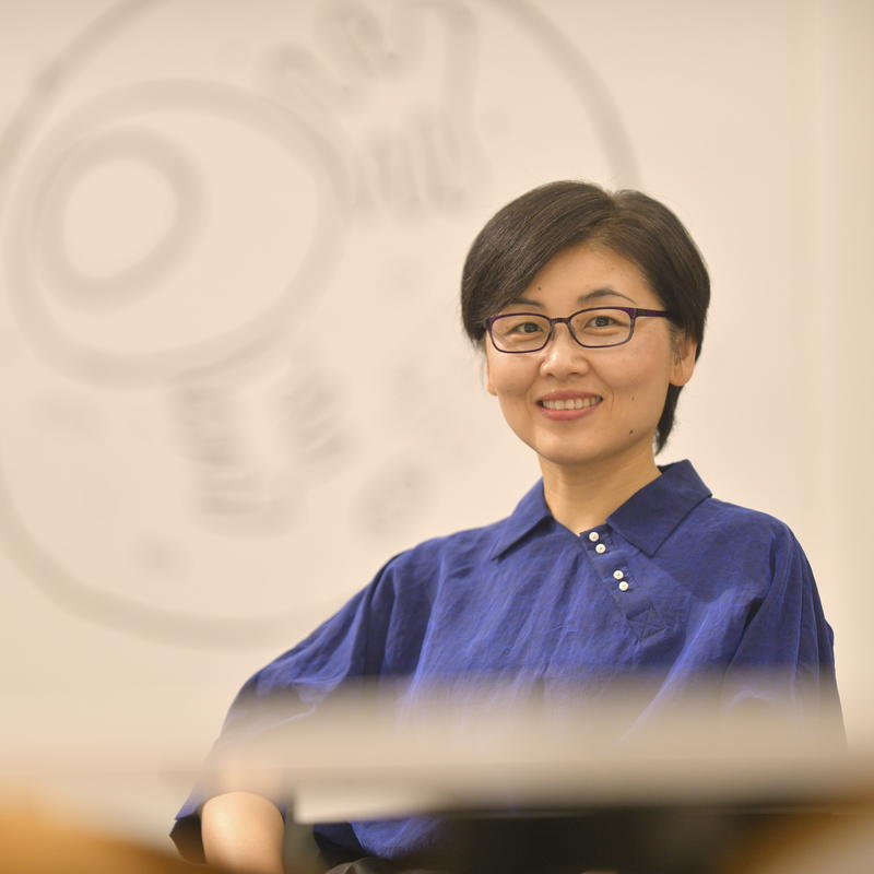 A woman with short dark hair, wearing an indigo tunic and glasses, smiles into the camera.