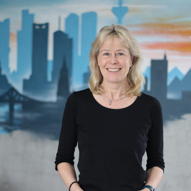 A head and shoulders of a woman smilinginto the camera, in front of a wall mural showing a city skyline of building silhouettes in a range of blue shades.