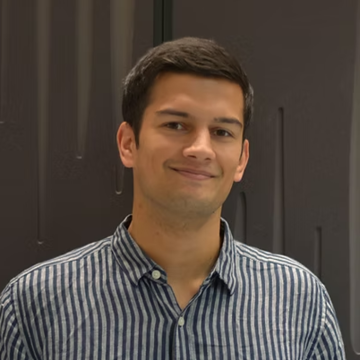 A young man in a blue-and-white striped shirt stands smiling in front of a dark wall
