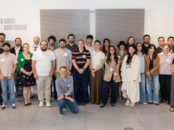 A large group of symposium attendees pose for a group photo inside the Fred Kavli Auditorium, standing in rows in front of a light-colored wall and auditorium signage.